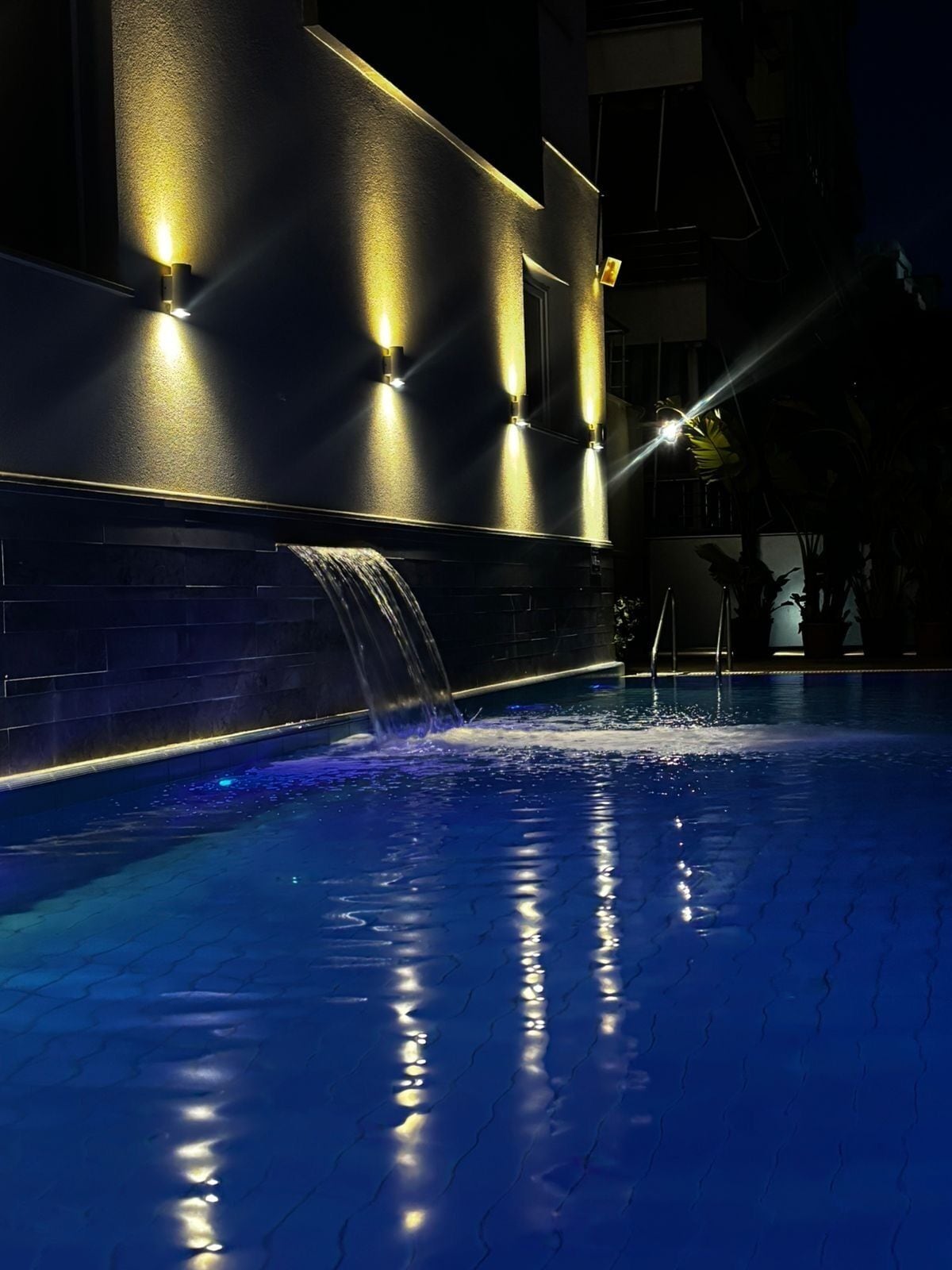 A swimming pool with a waterfall feature, illuminated by wall-mounted lights, and a dark surrounding area.