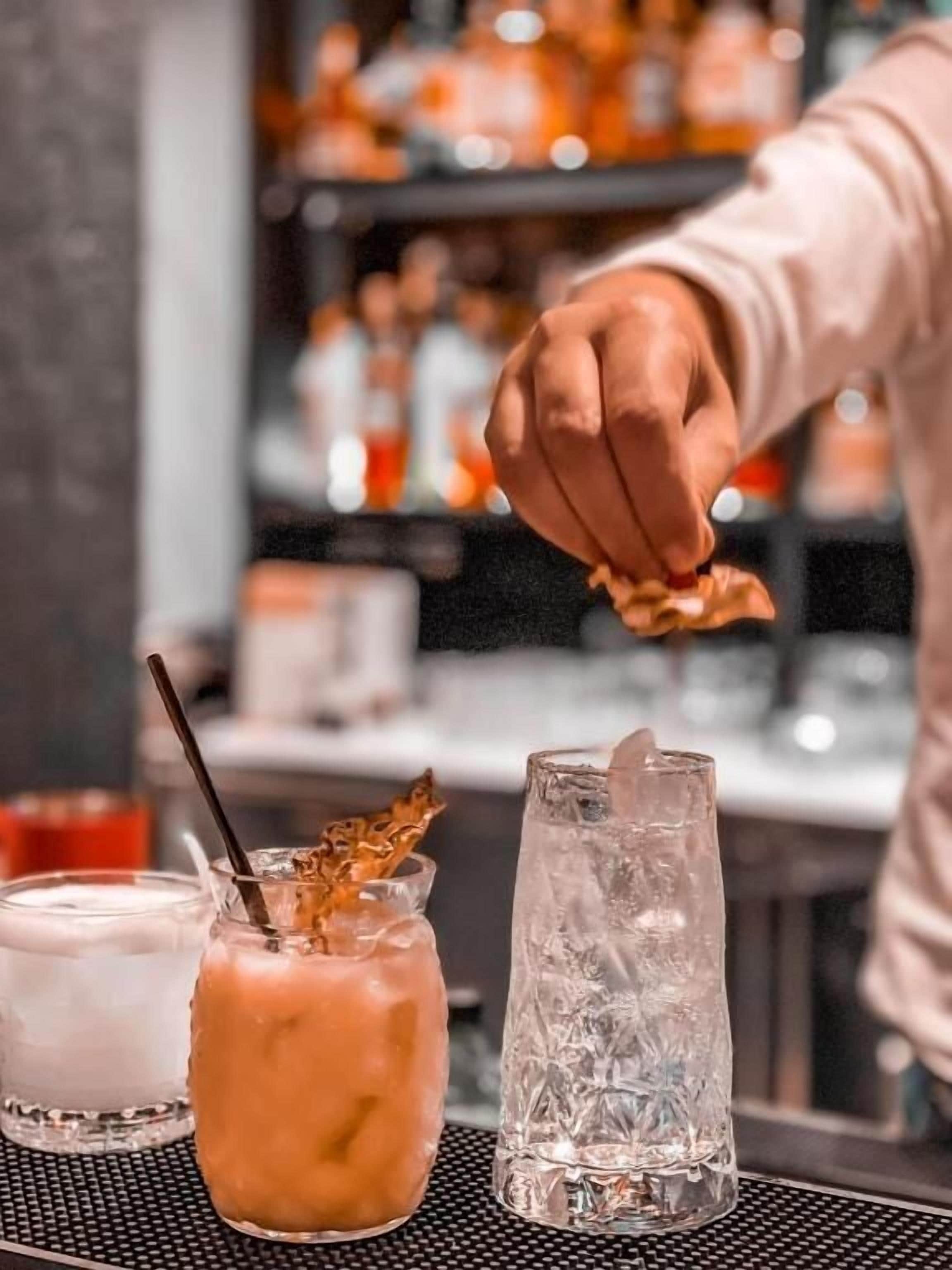 A bar setting with a bartender preparing a drink. There are three glasses on the counter: one with a straw and a decorative leaf, one with a creamy beverage, and one with ice cubes.