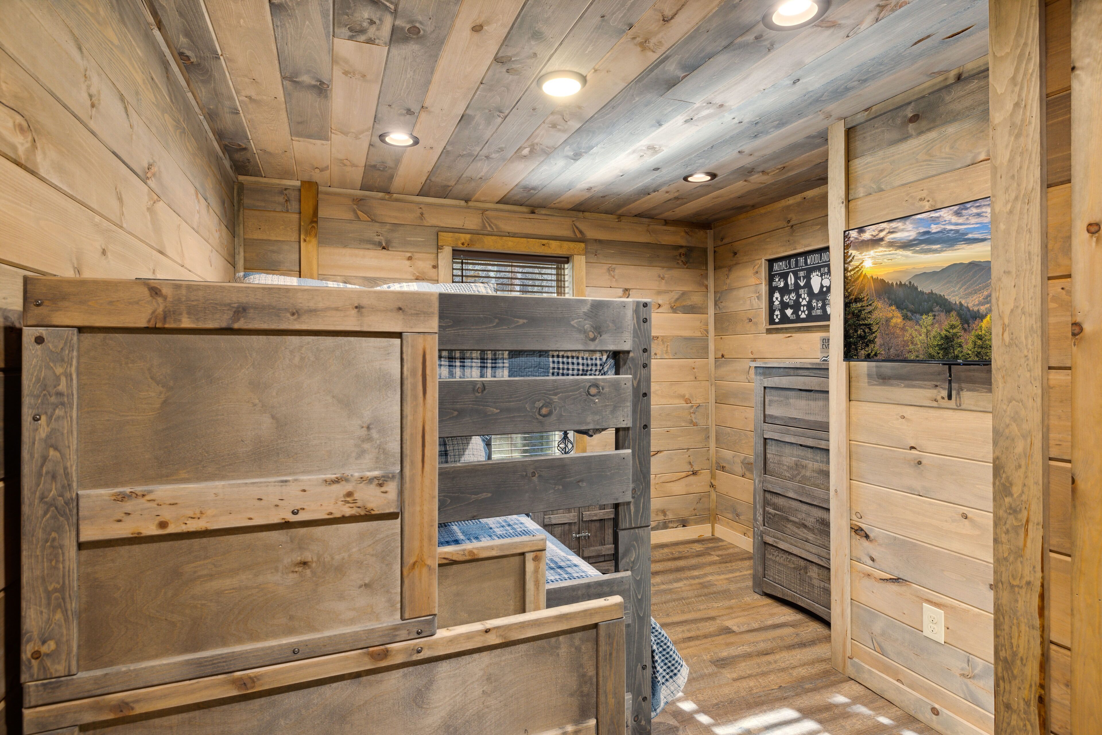 A wooden cabin interior with bunk beds, a window with blinds, and a scenic view poster on the wall.
