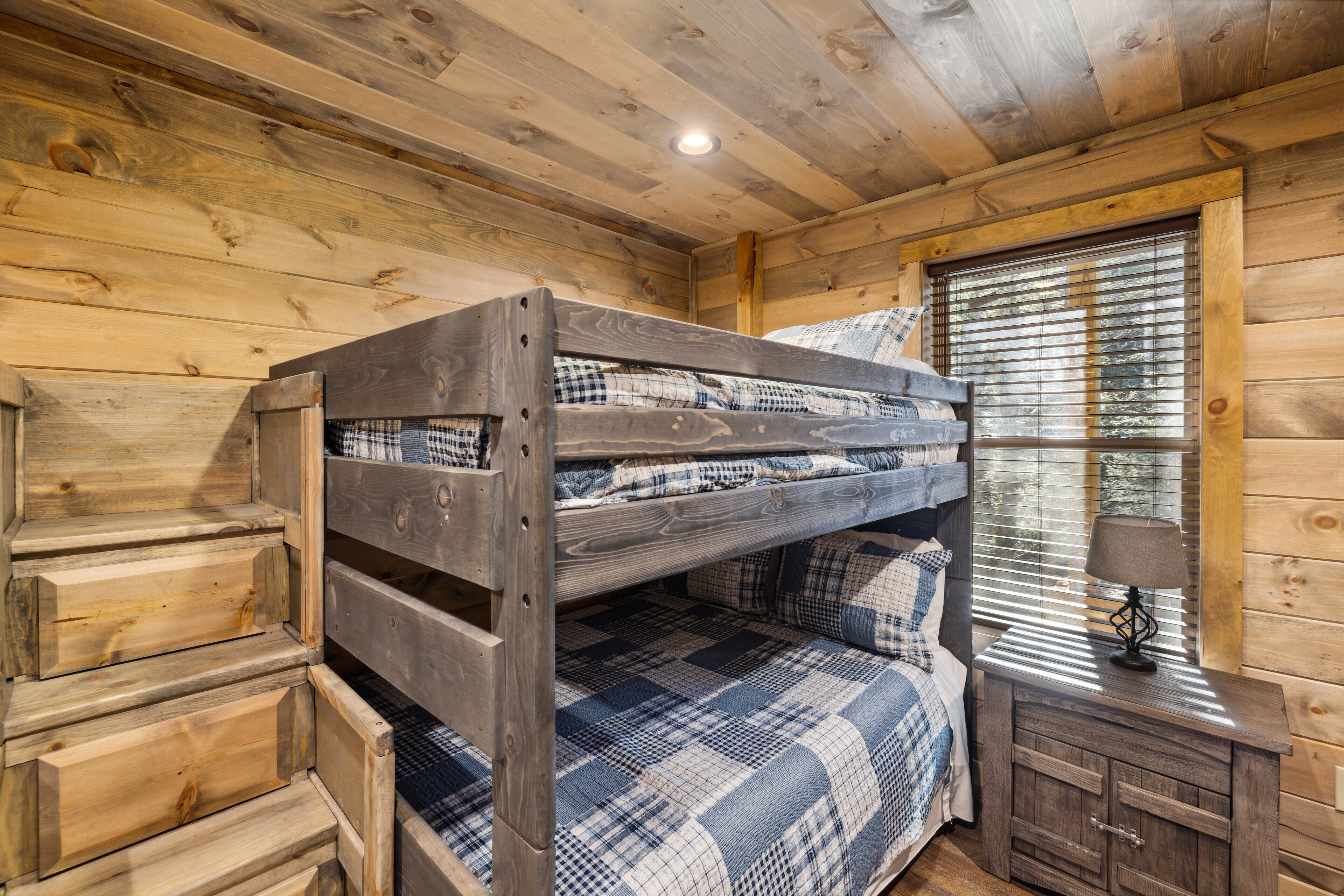 Una habitación tipo cabaña de madera con literas, una ventana con persianas y un pequeño armario de madera con una lámpara.