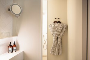 Une salle de bain avec un lavabo blanc, deux bouteilles de shampoing, un peignoir rayé accroché au mur et une pomme de douche.
