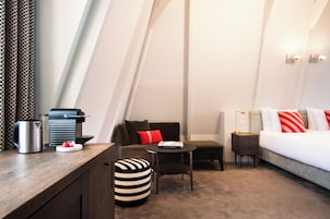 A modern hotel room with a cozy seating area, a small table, a sofa, a bed with red and white pillows, and a nightstand with a lamp.