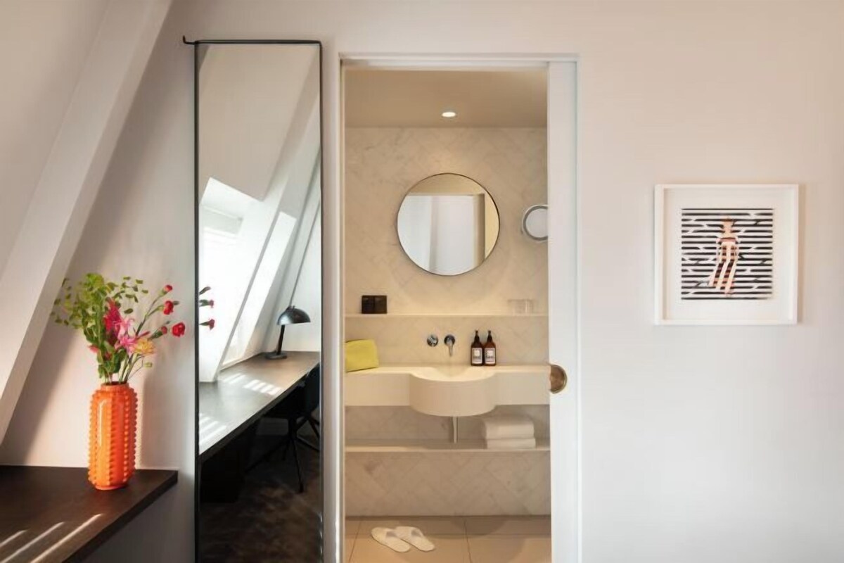 A modern bathroom with a round mirror, a sink, and a toilet. There is a framed artwork on the wall and a vase with flowers on the counter.
