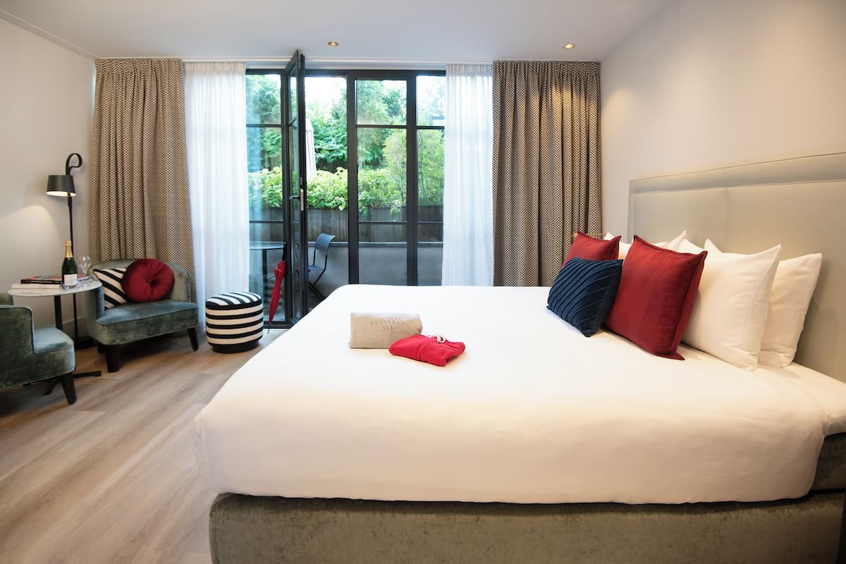 A modern hotel room with a large bed, a small seating area, and a balcony with a view of greenery.