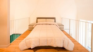 Uma cama de solteiro num quarto com teto inclinado, soalho de madeira e grades brancas.
