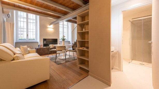 Uma sala de estar moderna com um sofá, uma mesa de centro e uma lareira. Há uma divisória de chuveiro de vidro e uma estante de livros.