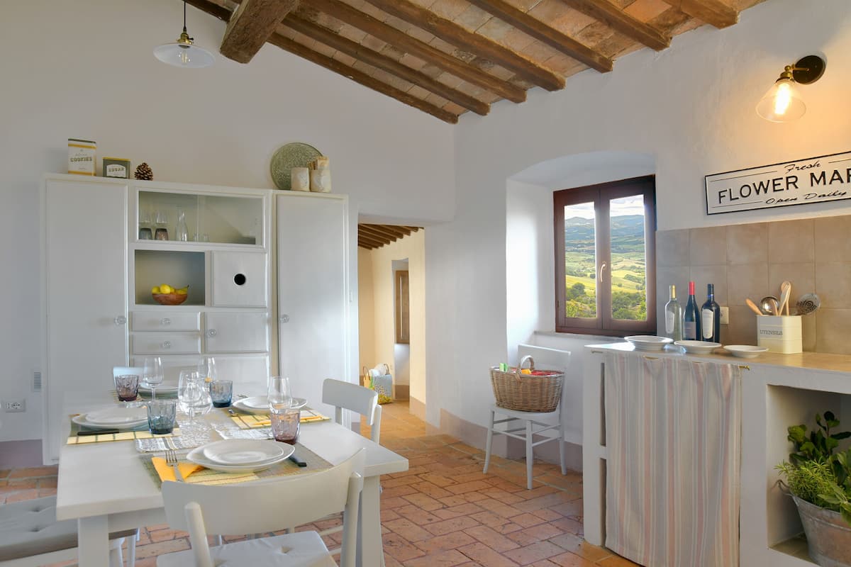 A well-lit kitchen with white cabinetry, a dining table set with plates and glasses, a window with a view of the countryside, and a sign indicating fresh flower delivery.