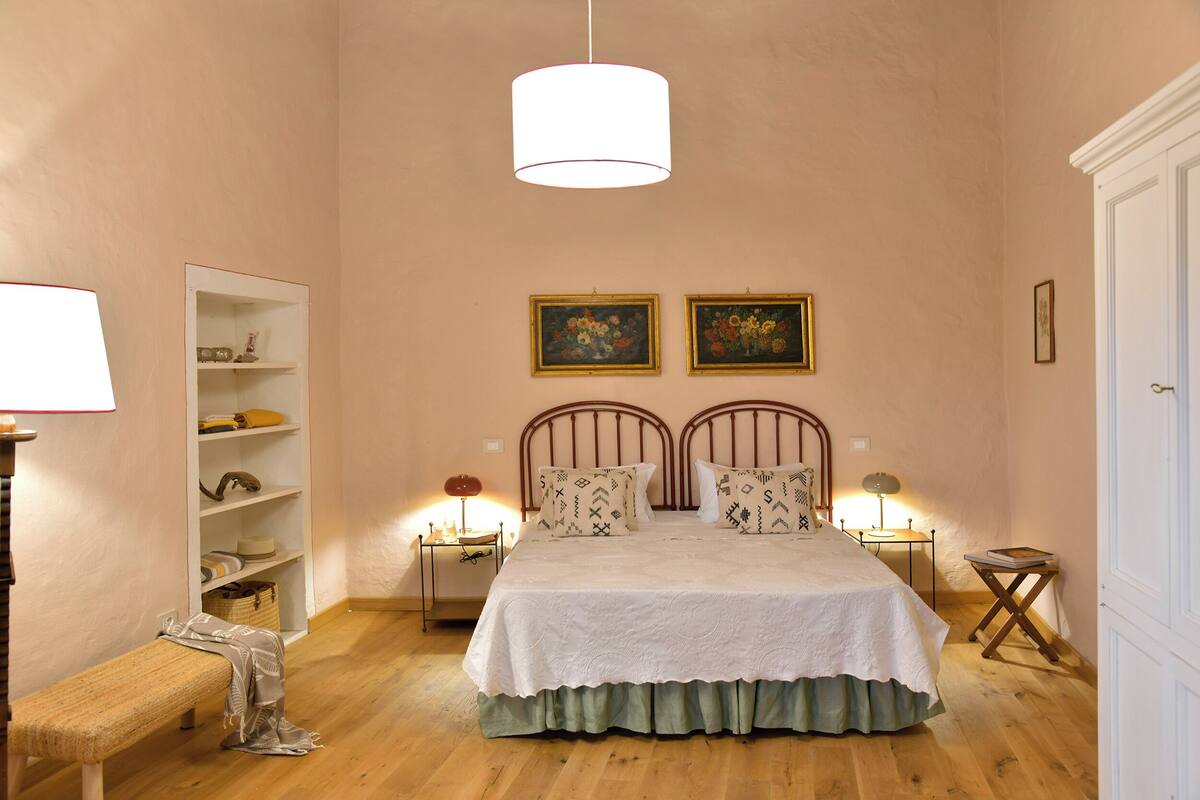 A bedroom with a bed, two bedside tables with lamps, a bench, a wardrobe, and two framed pictures on the wall.