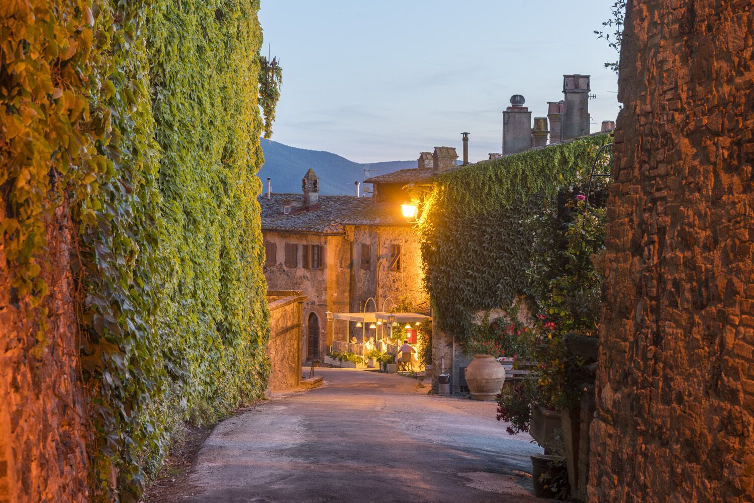A narrow alleyway with a building on the right side covered in ivy, a stone wall on the left, and a lit-up establishment in the distance.