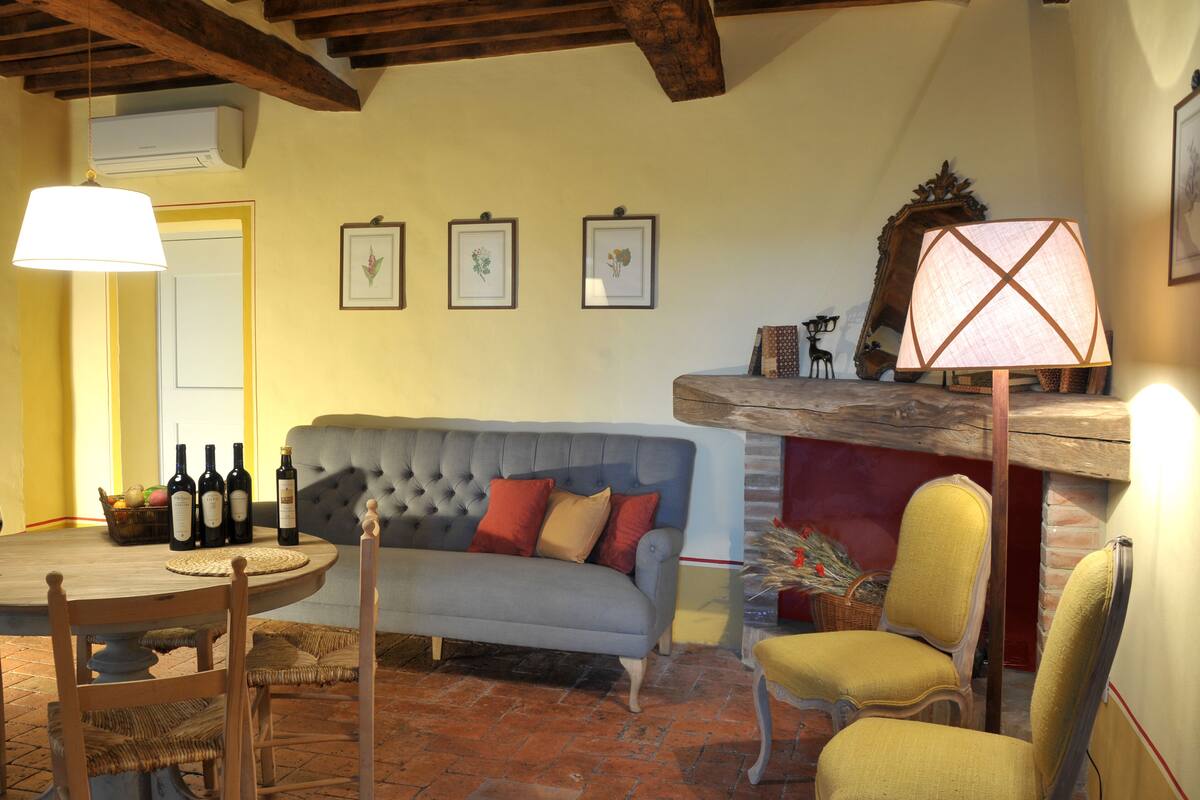A cozy living room with a sofa, chairs, a table with wine bottles, framed pictures on the wall, and a fireplace.