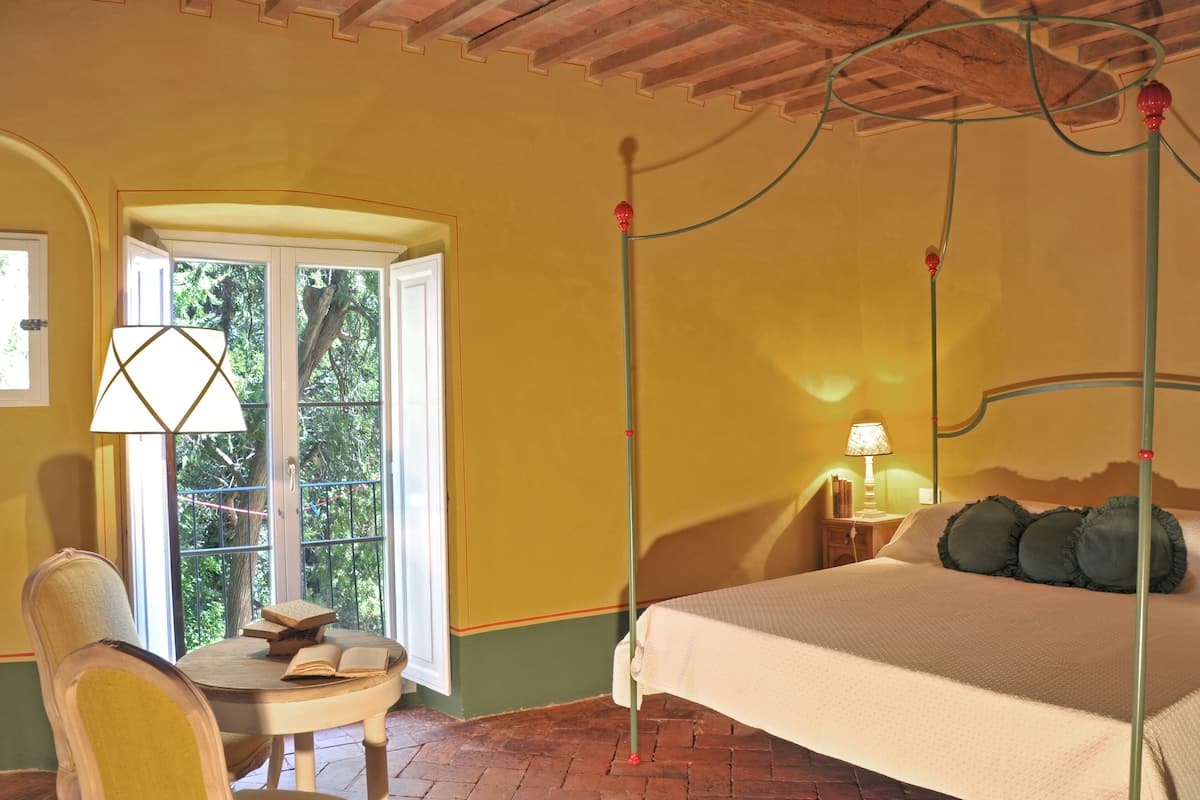 A bedroom with a four-poster bed, a chair, a small table with books, and a window with a view of greenery.