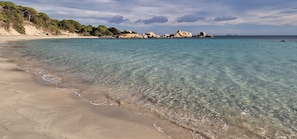 A beach with clear turquoise water, sandy shore, and rocky outcrops in the background.
