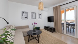Un salon moderne comprenant un canapé, une table basse ronde ornée d’une orchidée en pot, un téléviseur à écran plat et un balcon avec une table et des chaises pour prendre les repas.