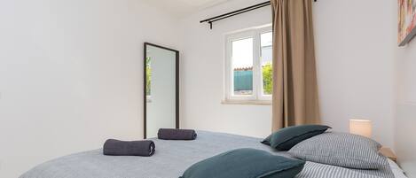 A bedroom with a bed, a window with curtains, a bedside lamp, and a wall-mounted mirror.