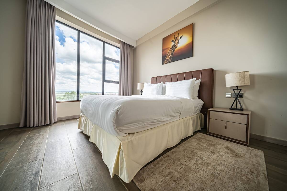 A hotel room with a large bed, a nightstand, a lamp, a painting, and a view of the sky.