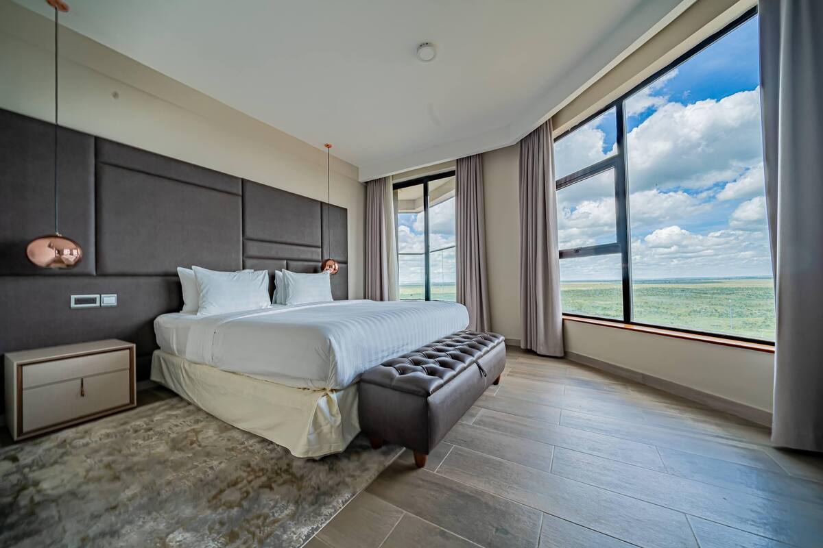 A modern bedroom with a large bed, a nightstand, a bench, and a view of the sky and landscape through a large window.