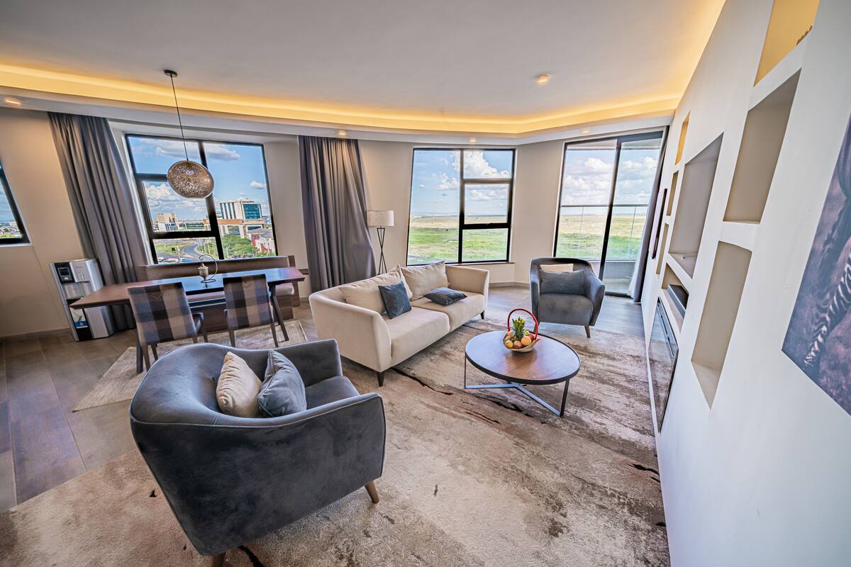 A modern living room with a sofa, armchairs, a coffee table, and a dining area with a table and chairs. Large windows offer a view of the cityscape.