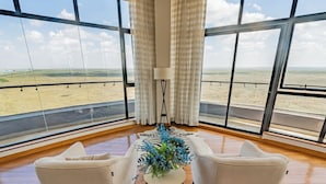 Amplia habitación con ventanas grandes que ofrecen vistas a un paisaje llano. Hay dos sillones blancos y una mesita redonda con un arreglo floral azul.