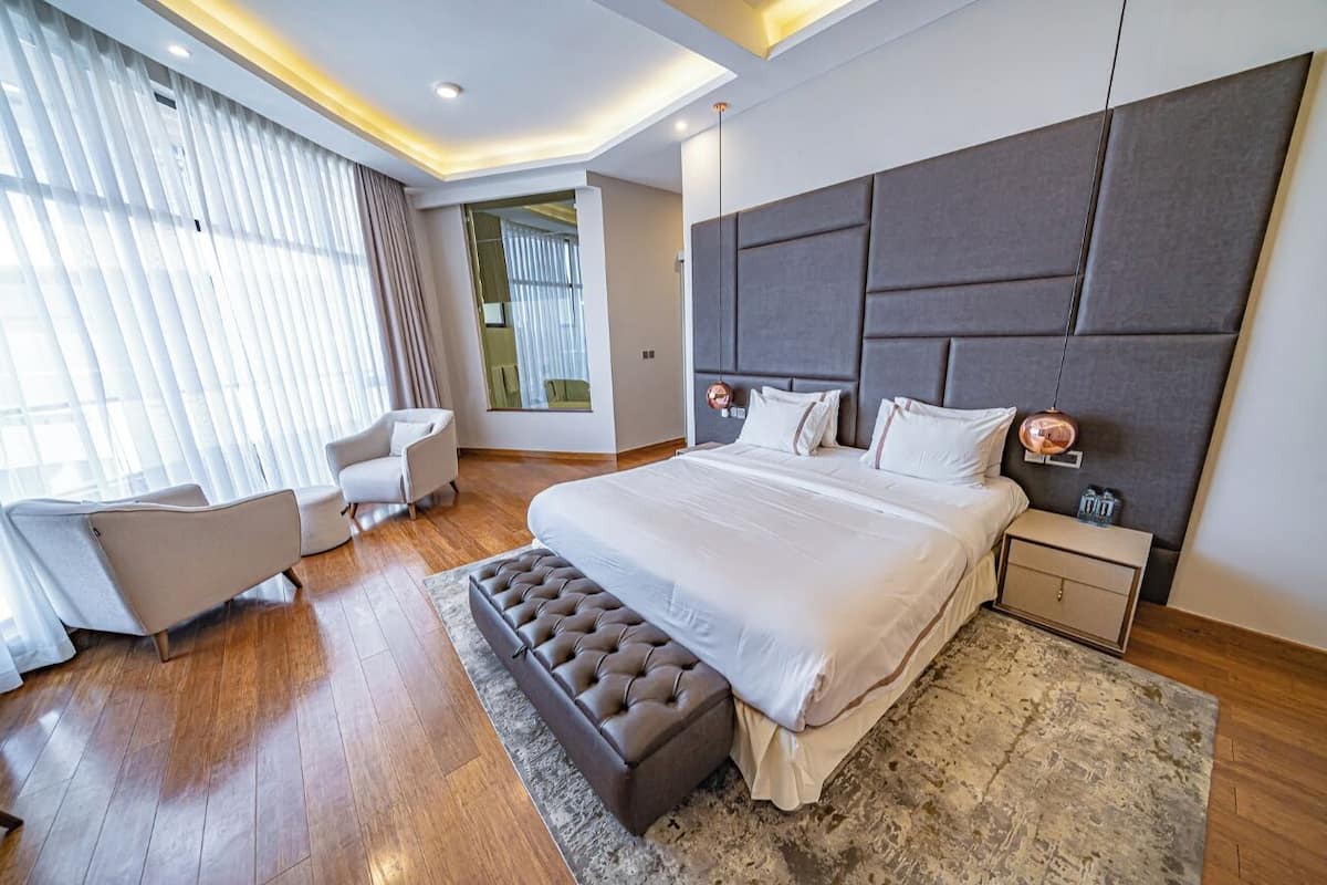 A modern hotel room with a large bed, a sitting area with a chair, and a view through a window with sheer curtains.