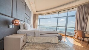 Un dormitorio moderno con una cama grande, una mesita de noche, una silla y vistas de la ciudad a través de amplias ventanas.
