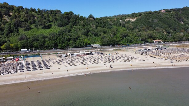 A beach with numerous umbrellas and sun loungers, a lifeguard tower, and a building with a red roof in the background.