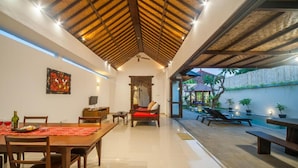 A spacious indoor area with a dining table, chairs, a sofa, and a poolside seating area with lounge chairs and a small table.
