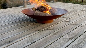 Een vuurplaats met houtblokken en dennenappels op een houten terras met uitzicht op een waterpartij.