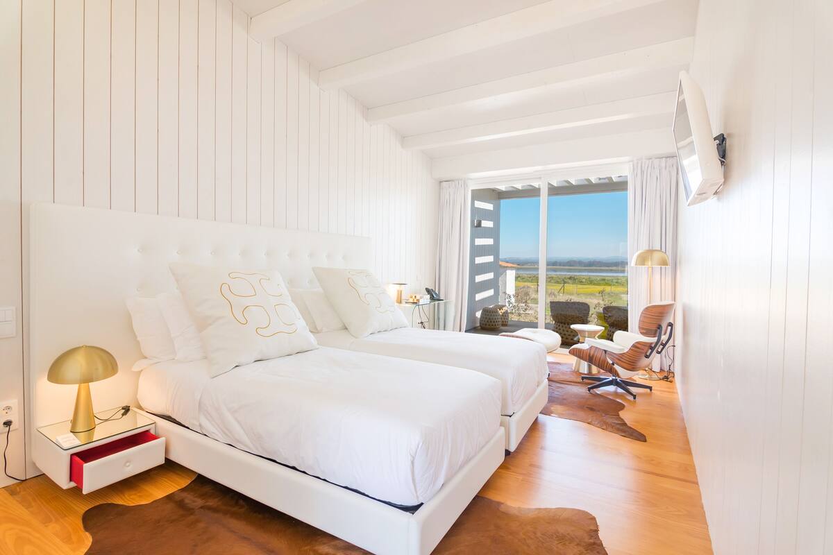 A bedroom with two beds, a nightstand, a lamp, and a view of the outdoors.