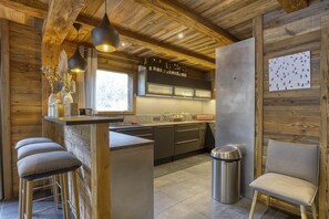 A modern kitchen with wooden accents, bar stools, a trash can, and a chair.