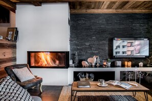 A cozy living room with a fireplace, a comfortable sofa, a coffee table with books and a cup, and a TV mounted on a black brick wall.