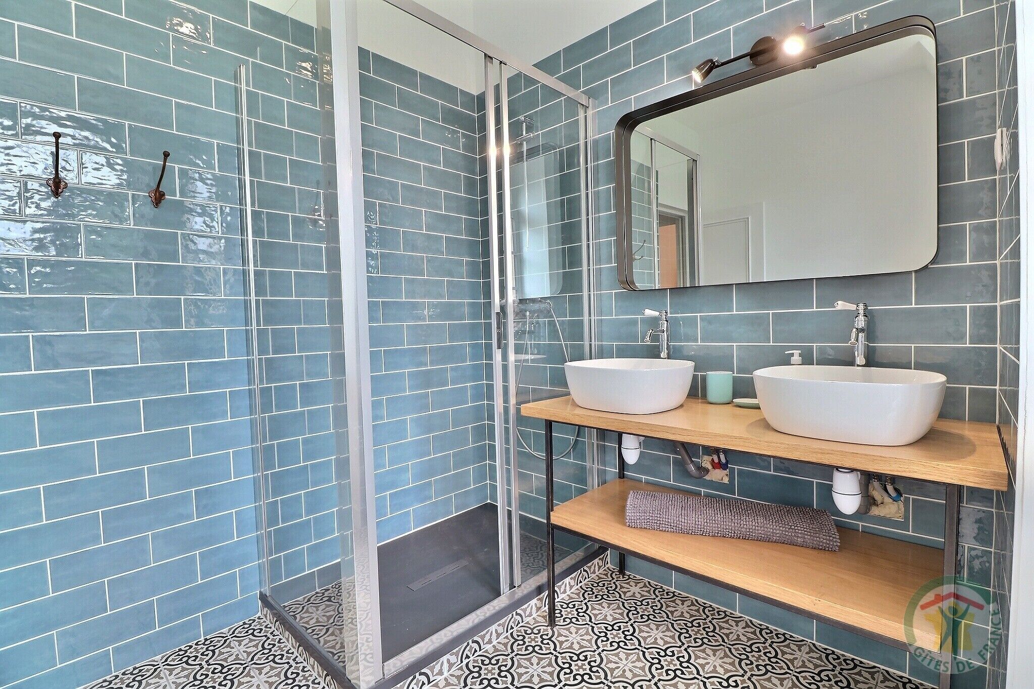 Ein Badezimmer mit blauen Fliesen, einer Duschkabine aus Glas, zwei Waschbecken auf Holzablagen und einem großen Spiegel darüber.