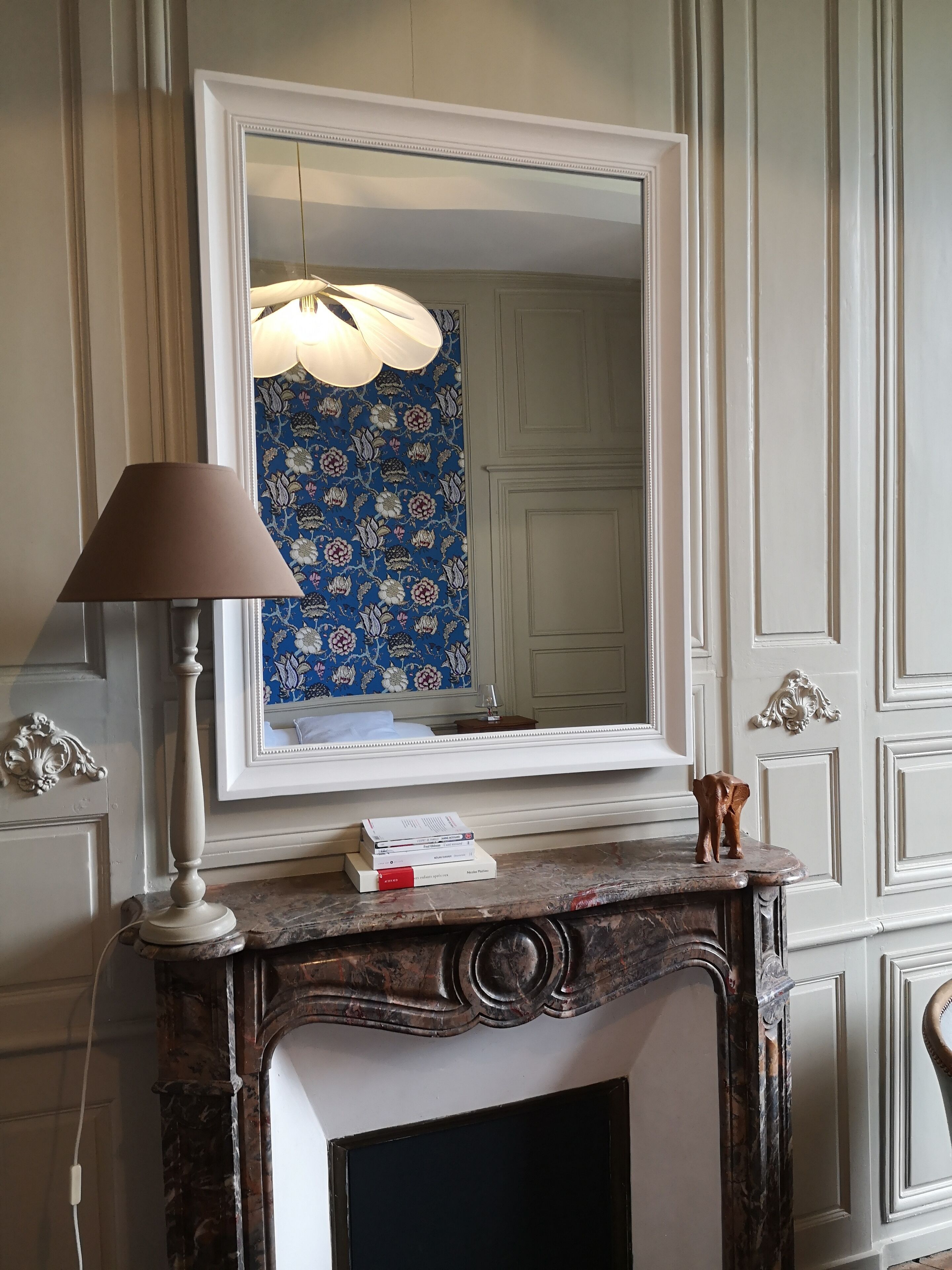 Een kamer met een grote spiegel, een lamp, een open haard en behang met een patroon.