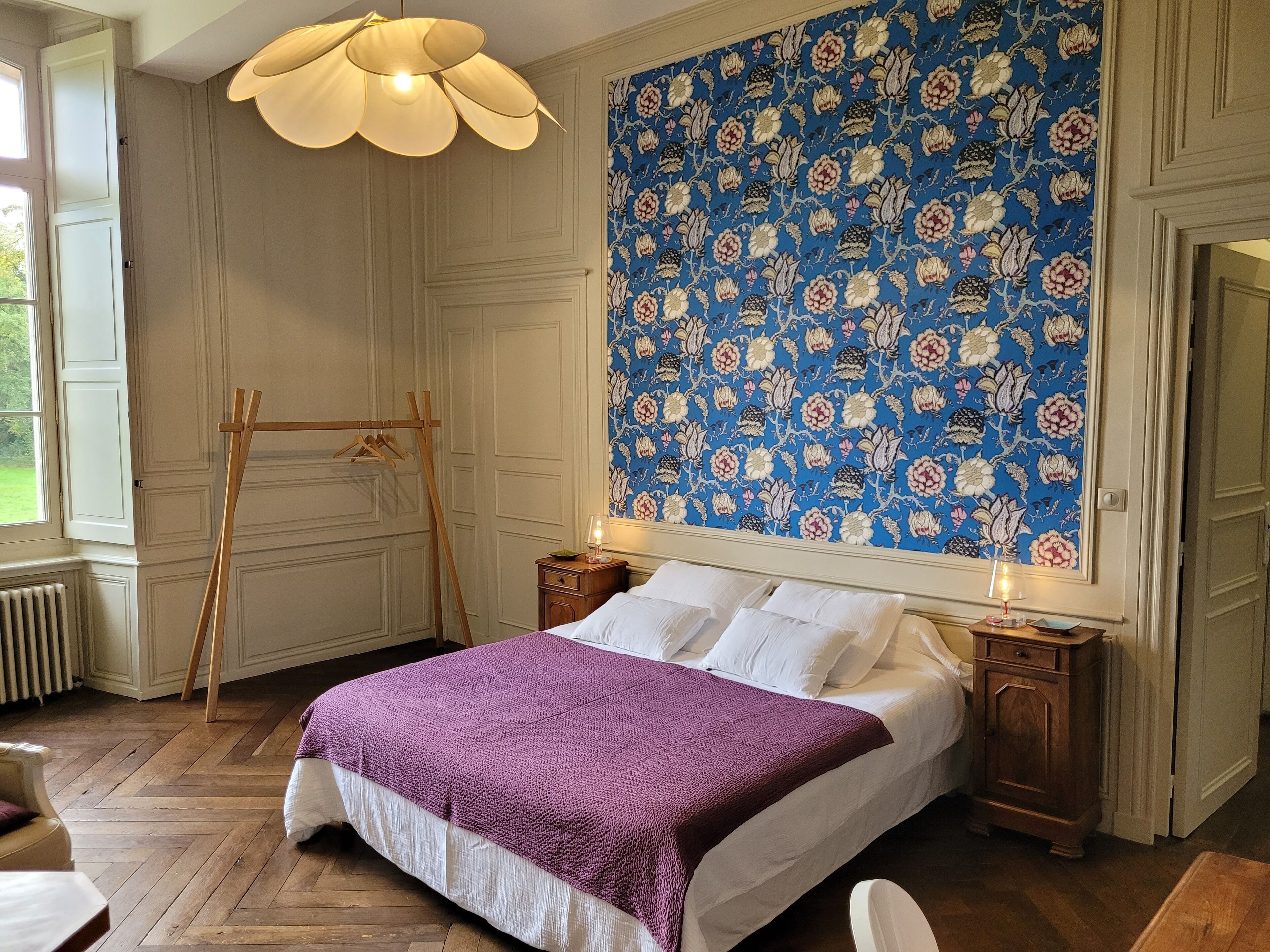 Un dormitorio con cama, cómoda de madera, mesita de noche, lámpara colgante y papel tapiz floral.