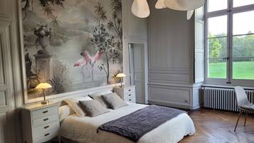 A bedroom with a large mural, a bed with multiple pillows, bedside tables with lamps, and a view through a large window.