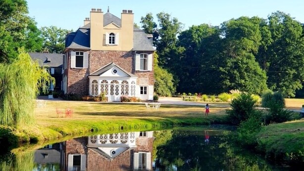 A large, two-story house with a stone facade and a large front porch, surrounded by lush greenery and a calm pond. - Domaine des Longrais (Saint-Thurial)