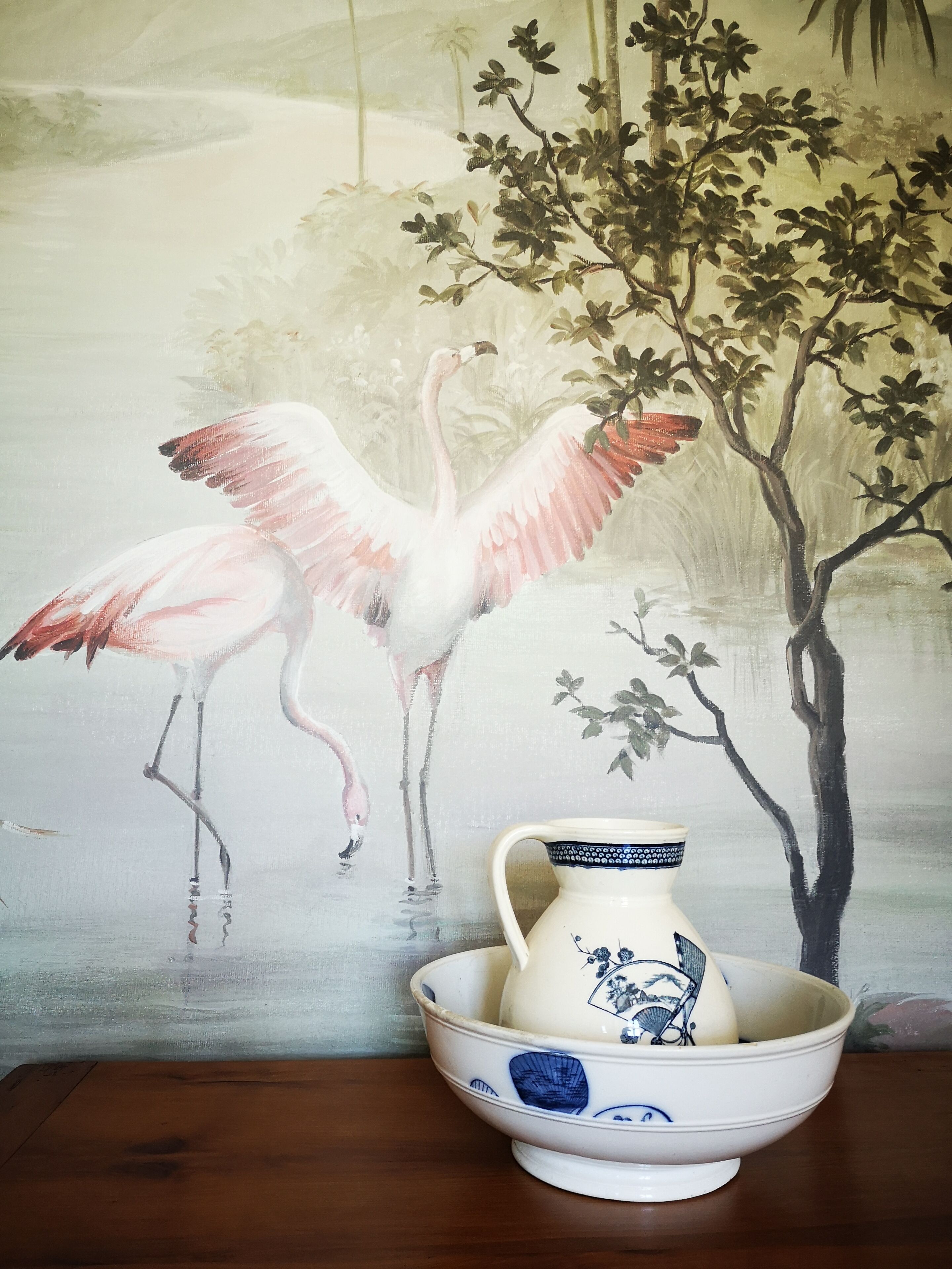 Een schilderij van flamingo's en een boom op een muur, met een witte keramieken kan op een houten tafel ernaast.