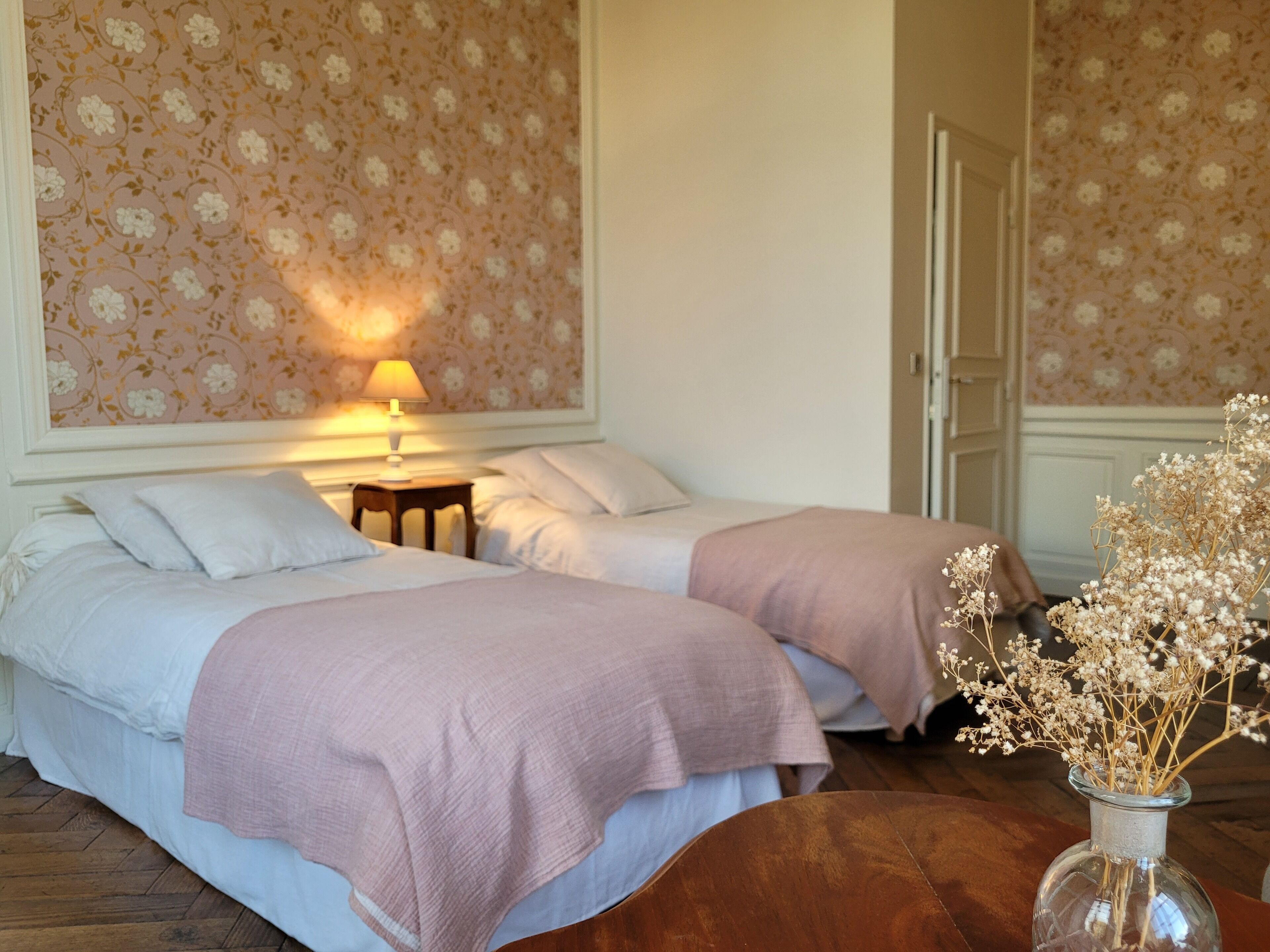 Quarto de hotel com duas camas, uma mesa de cabeceira, um abajur e um vaso com flores secas.