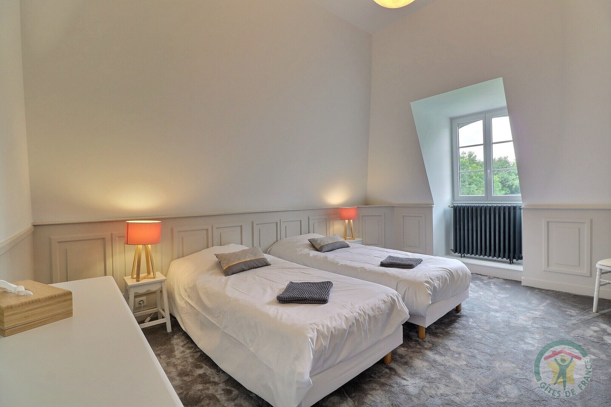 A bedroom with two beds, a nightstand with a lamp, a radiator, and a window with a view.
