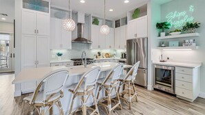 A modern kitchen with white cabinetry, a central island with bar stools, stainless steel appliances, and a neon sign on the wall. - Designer Interiors@Cinnamon Shore in Beach Home w/ Resort-Like Amenities! (Port Aransas)
