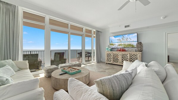A spacious living room with a large sliding glass door leading to a balcony, a flat-screen TV mounted on the wall, and a wooden coffee table.