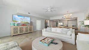 A spacious living room with a large TV, a white sofa, a wooden coffee table, and a kitchen area in the background.