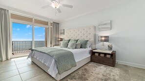 A bedroom with a large bed, a nightstand, a ceiling fan, and a view of the sea through large windows.