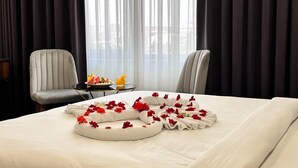 Une chambre d’hôtel avec un lit bien fait, une petite table sur laquelle se trouve un panier de fruits et des boissons, et deux chaises.