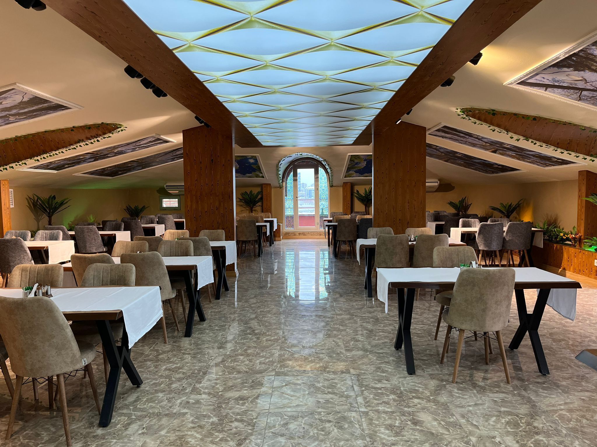 A spacious dining area with multiple tables and chairs, a high ceiling with a geometric pattern, and a view through an arched doorway.
