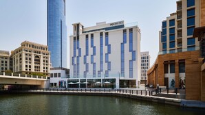 A modern hotel with a distinctive zigzag facade, a glass skyscraper to the left, and a river in the foreground. - INK Hotel (Dubai)