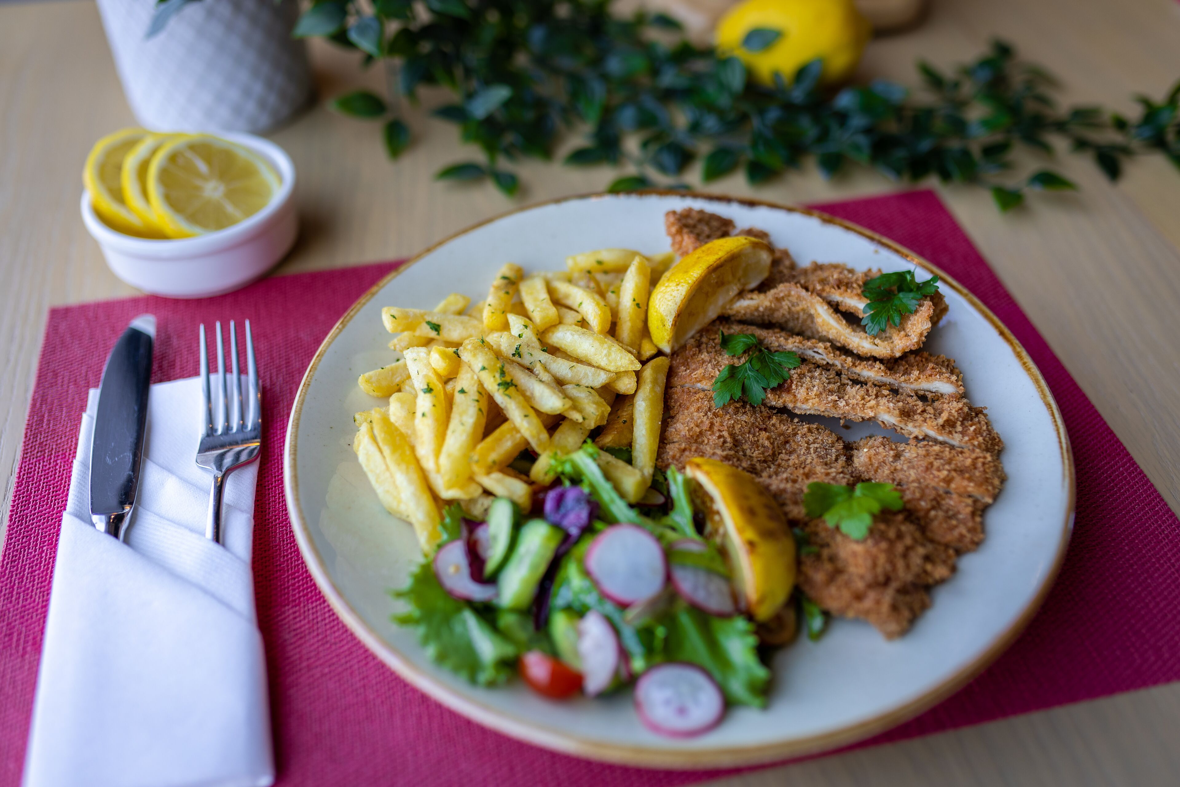 Un piatto di cibo con patatine fritte, un'insalata e merluzzo impanato, accompagnato da fette di limone e una piccola ciotola con spicchi di limone.