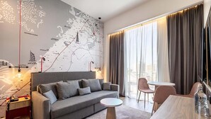 A modern hotel room with a grey sofa, a round coffee table, a dining table, and a large window with curtains. - INK Hotel (Dubai)