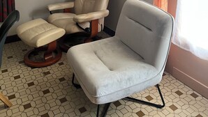 Une pièce comprenant un fauteuil en cuir beige, un pouf assorti, un fauteuil gris, un sol carrelé, des tableaux encadrés accrochés au mur, un vase posé sur une étagère et une fenêtre ornée de rideaux orange.