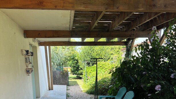 Un patio couvert avec une table et des chaises, un chemin en béton et un jardin agrémenté de plantes et de fleurs.