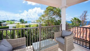 Un balcon doté de meubles en osier et d’une table en verre, offrant une vue sur une zone côtière avec des maisons et des arbres.